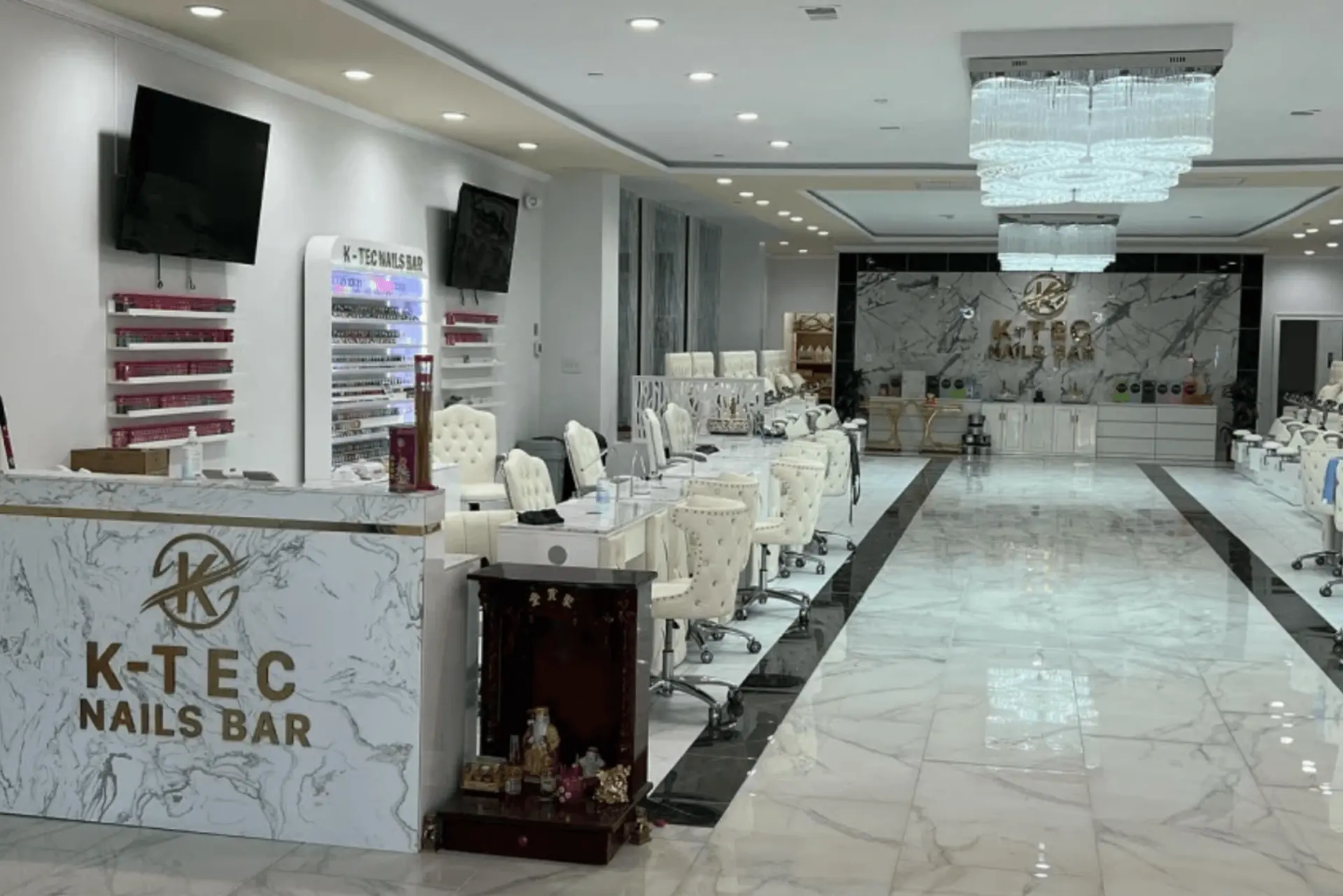 A spacious, modern nail salon named "K-TEC NAILS BAR" with white marble floors and counters. The salon features multiple manicure stations with cream-colored chairs, pedicure chairs in the background, and large chandeliers. The front desk is white marble with a gold logo. There's a display of nail polishes and two TVs on the left wall.